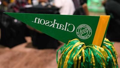 A green pennant with Clarkson University's logo is displayed nestled into a green and yellow pom-pom